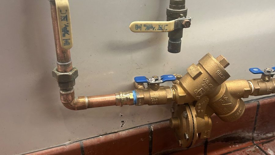 A brass backflow prevention valve assembly connected to copper plumbing pipes near a tiled floor, with multiple shutoff valves attached.