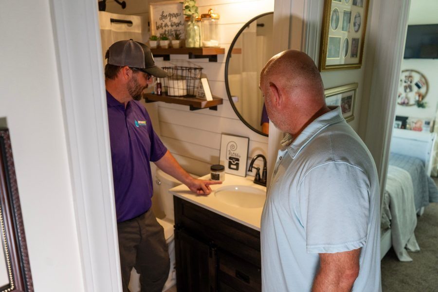 Two men in a home bathroom: a repair technician in a purple shirt explains something at a white sink while a homeowner watches.