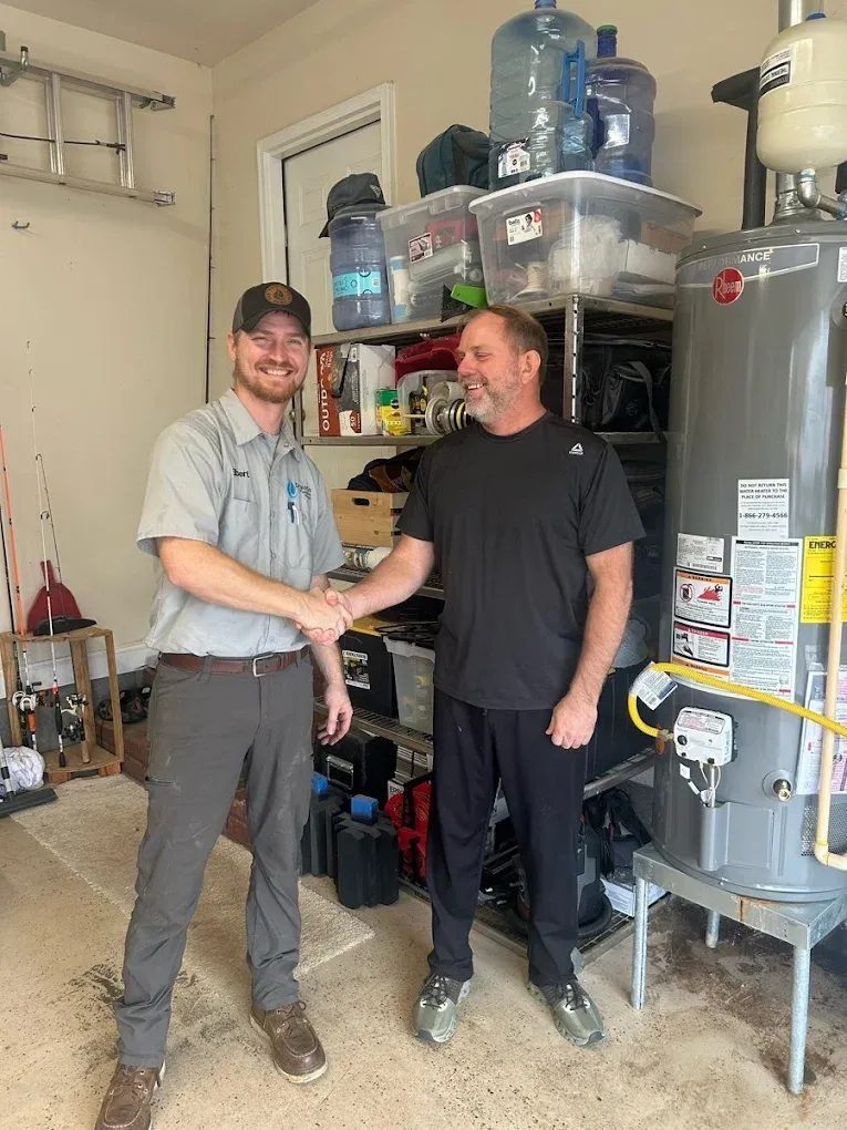 Two men shaking hands in a garage or utility room, standing beside a large water heater and storage shelves.