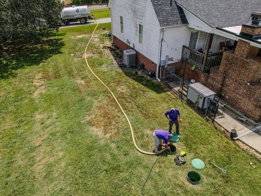 Aerial view of two workers in purple shirts handling a long hose that runs from a street tanker to an open green utility pit in a grassy backyard near a white house.