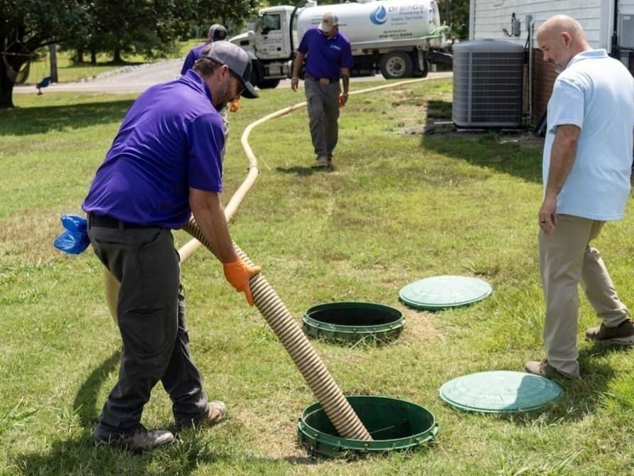 Maintenance crew in purple shirts handles a large hose into an open green underground access in a grassy yard, with a vacuum truck in the background.