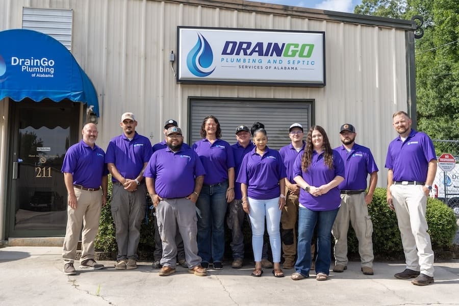 Group of DrainGo Plumbing employees posing outside their building in purple polo shirts with the company sign above.
