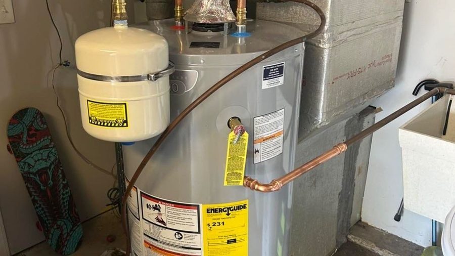 A residential water heater installed next to an HVAC system, with an expansion tank mounted above and copper piping connected to the unit.