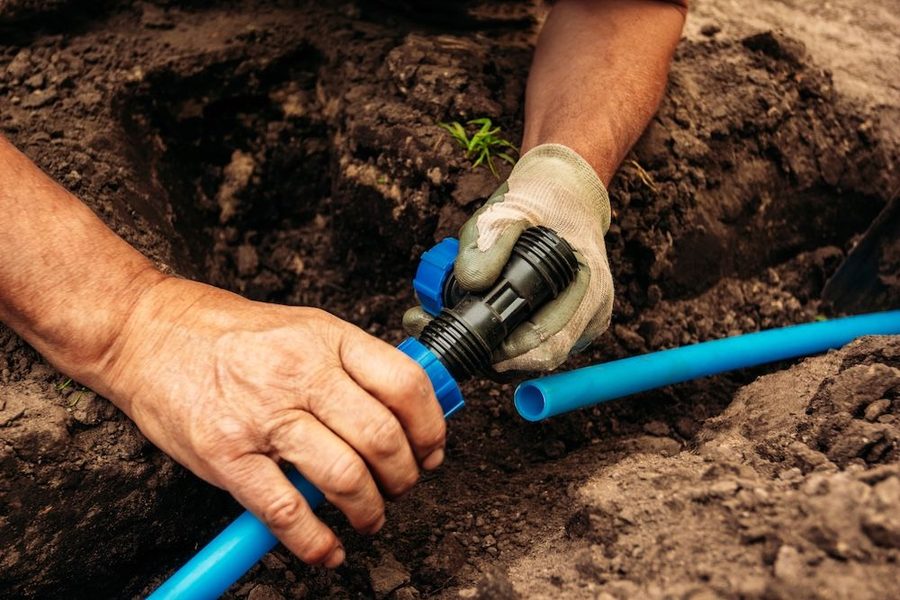 Close-up of gloved hands connecting a blue plastic irrigation pipe with a black connector in dirt in a trench.