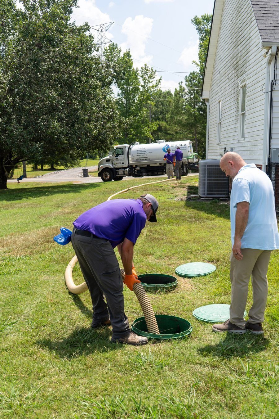 Workers insert a large hose into an open green manhole in a grassy yard, with a service truck and two men in the background.