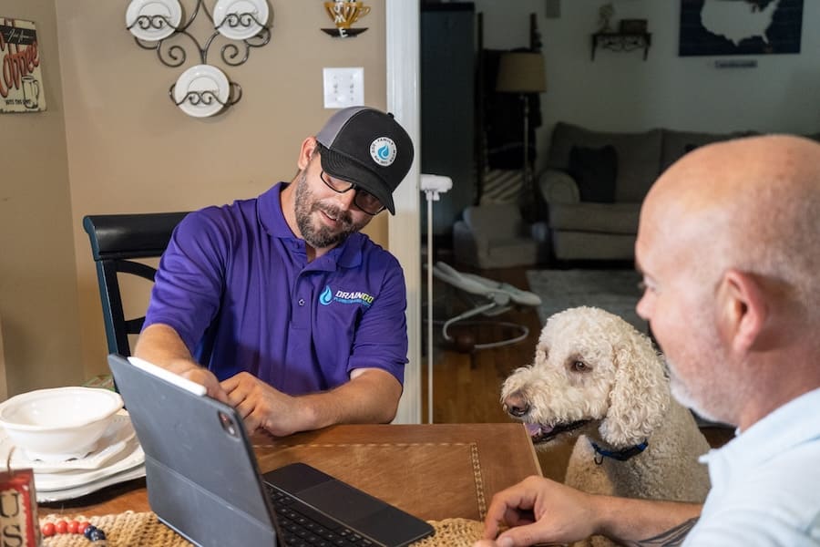 Plumber in a purple polo and cap shows a tablet to a client at a dining table, with a cream poodle nearby in a home setting.