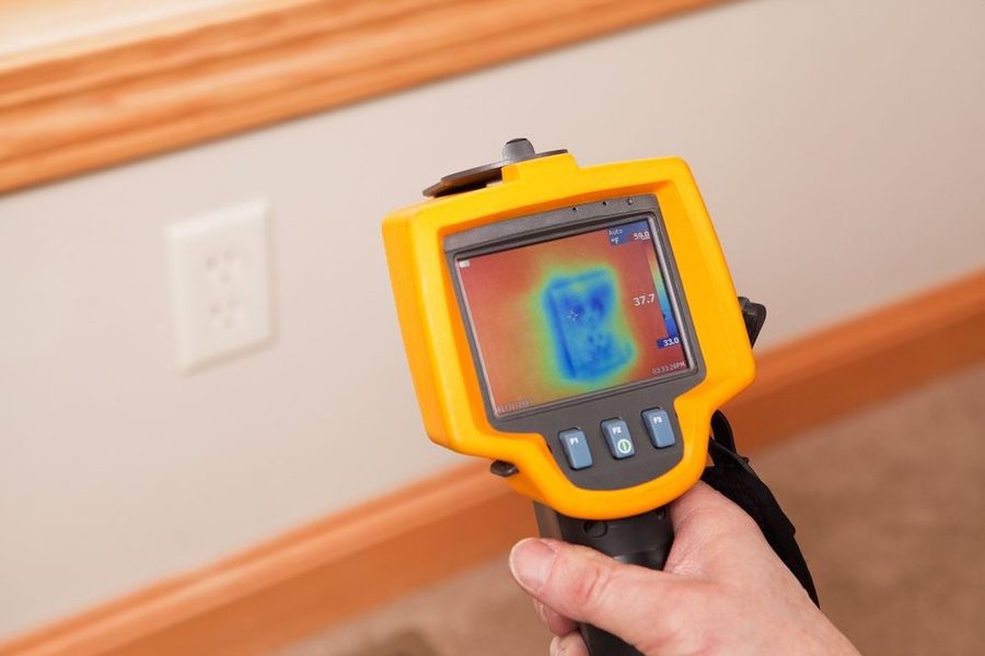 Handheld infrared thermal camera displaying a heat map of a wall near the floor/baseboard edge.