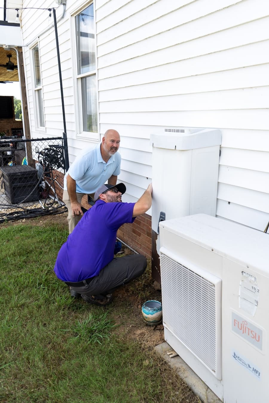 Two men installing or repairing an outdoor air conditioner unit beside a white house.