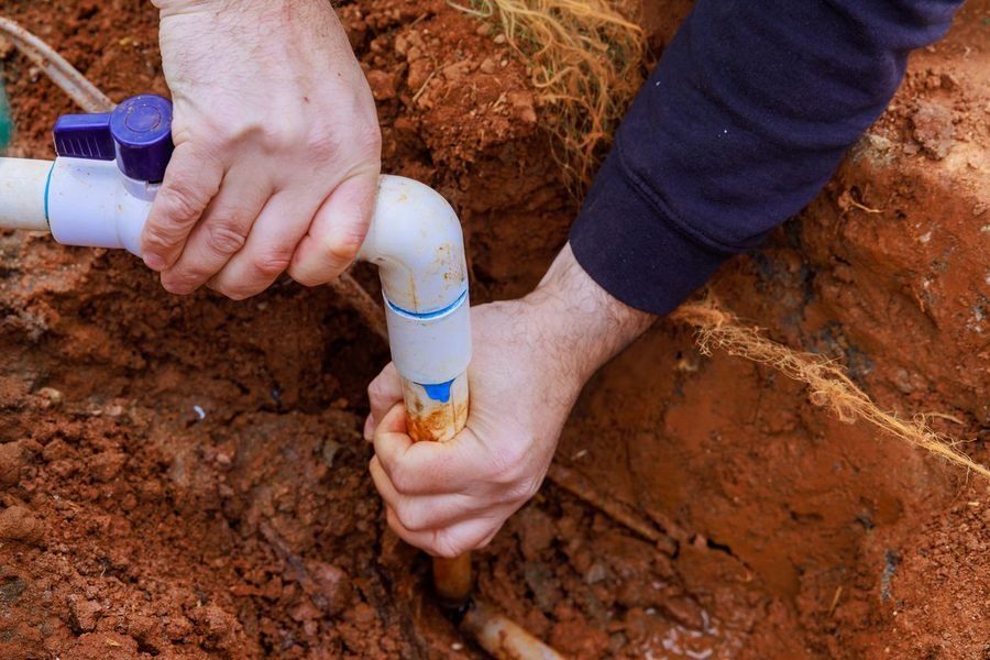 Close-up of hands assembling a white PVC pipe in a dirt trench during a plumbing repair.