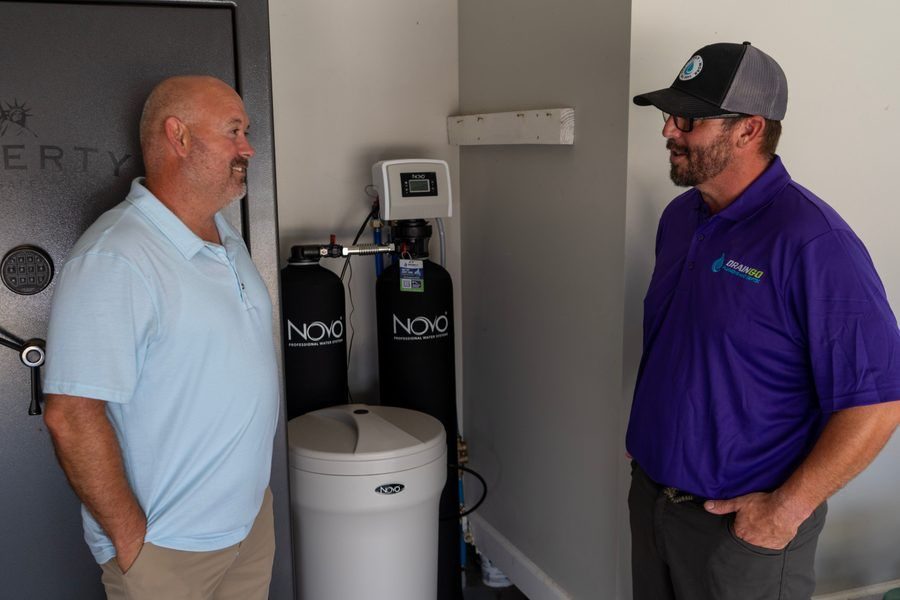Two men stand in a utility room talking beside a Novo water softener system with a white storage tank and digital control panel.