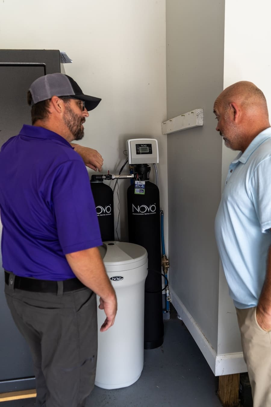 Two men inspect a Novo water filtration system with a white storage tank and black filter cylinders in a utility room.